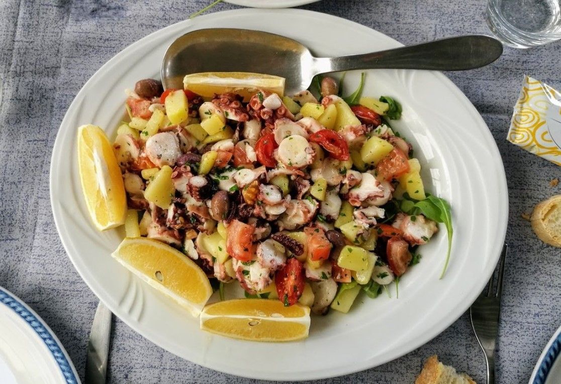 Insalata di polpo con patate, pomodori, olive, fette di limone e cucchiaio da portata su un piatto bianco.