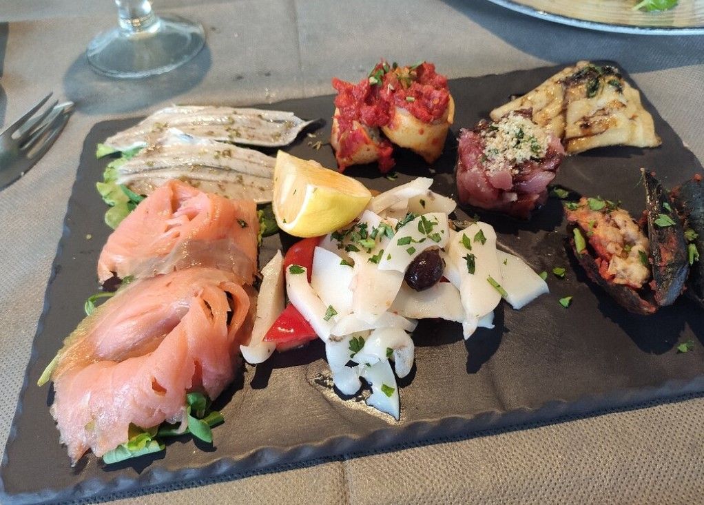 Piatto di antipasti di mare su ardesia nera: salmone, pesce bianco, olive e verdure grigliate.