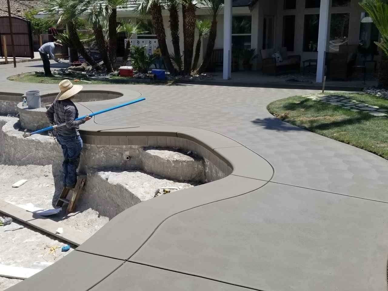 Worker smoothing fresh concrete around a curved patio edge beside a tree-lined building entrance