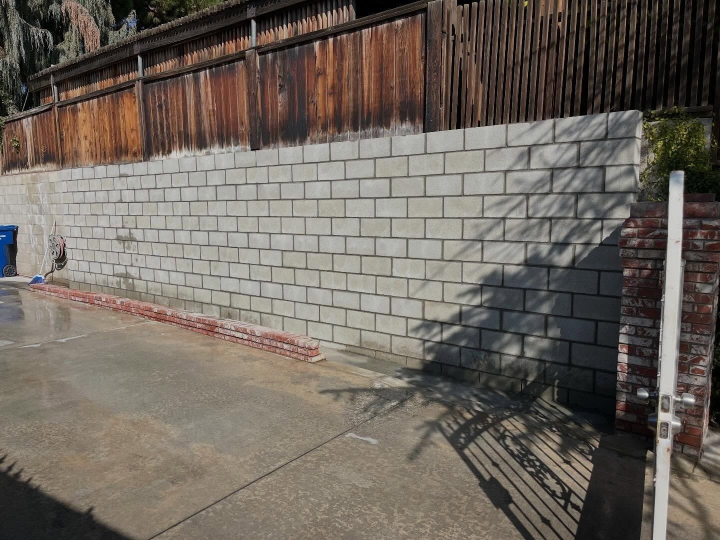 Concrete block retaining wall beside a wet sidewalk, with wooden fence above and deep shadow on the right.