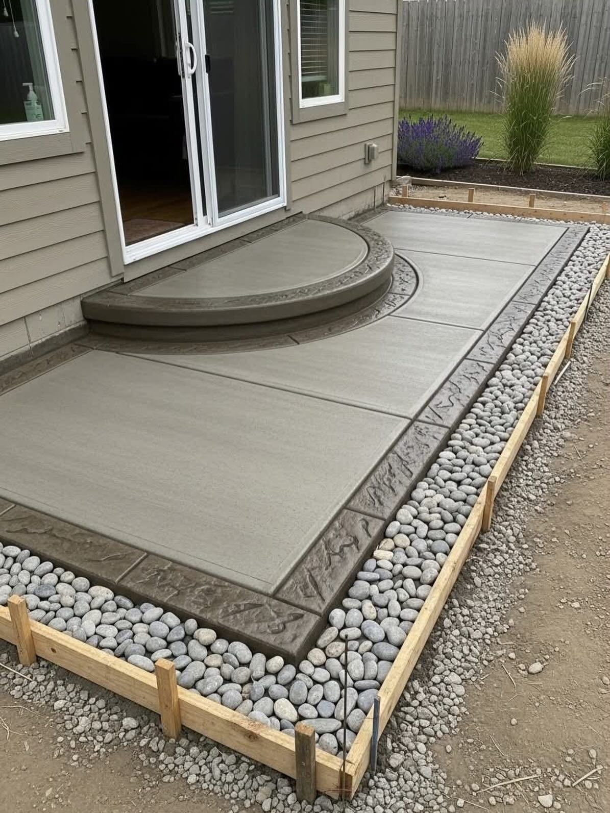 Raised concrete patio with rounded step, bordered by river rocks and wooden edging beside a house.