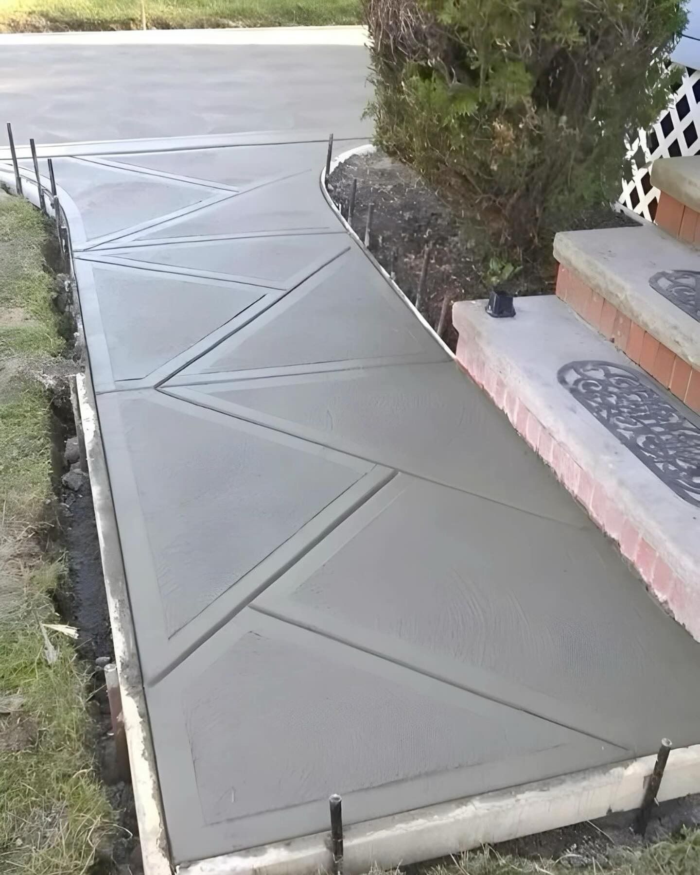 Concrete sidewalk with geometric patterns beside house steps and shrubbery, leading to a driveway.