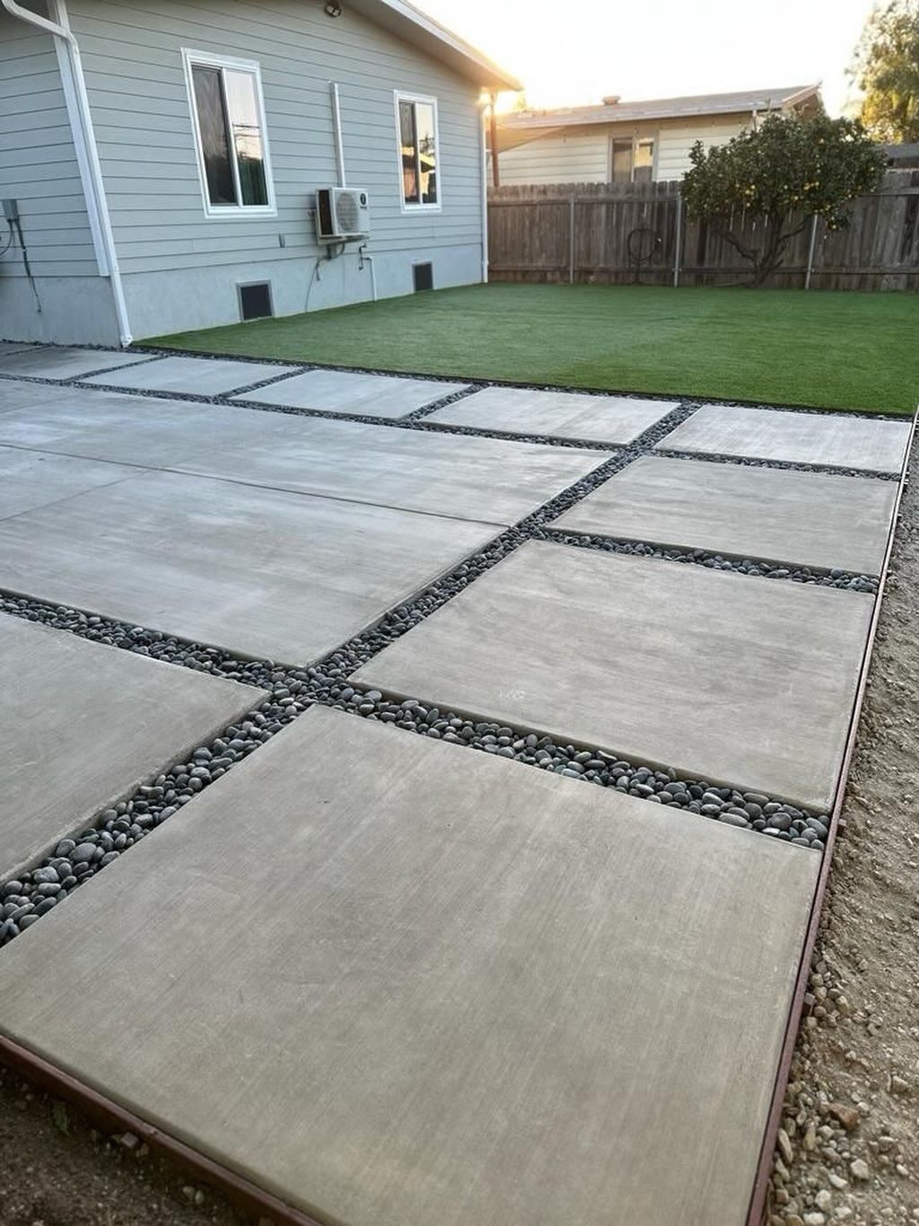 Backyard concrete patio with gravel joints beside a house and green lawn, fence in background