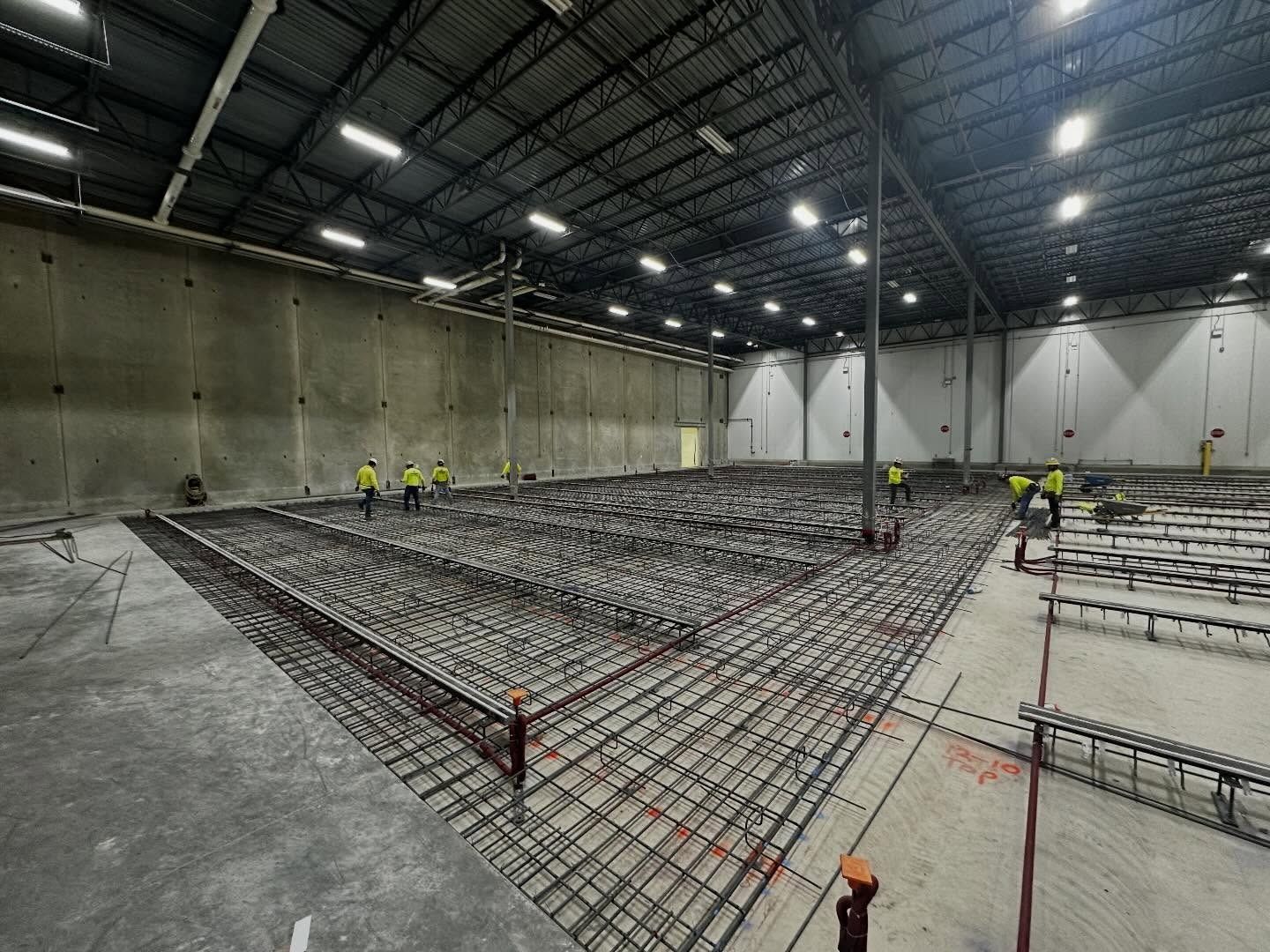 Workers installing rebar grid in a large industrial warehouse under bright overhead lights
