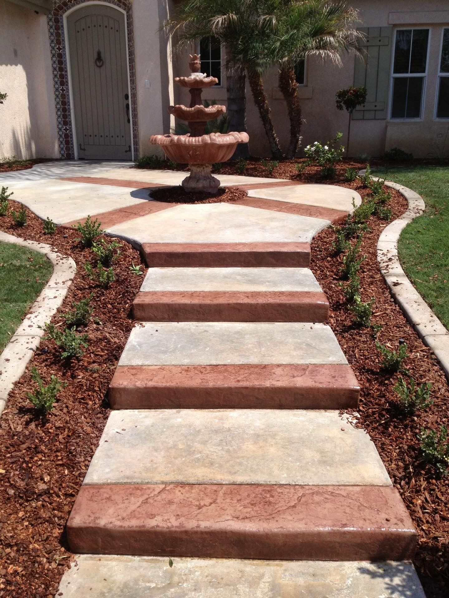 Front yard stone walkway leading to a tiered fountain, bordered by reddish gravel and shrubs.