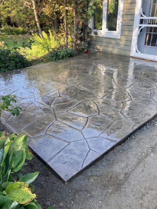 Stamped concrete patio beside a stone house, wet and reflective in sunlight, with garden plants nearby