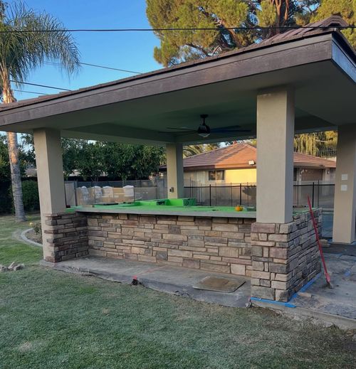 Stone outdoor pavilion with a green countertop, beige columns, and a dark roof in a yard.