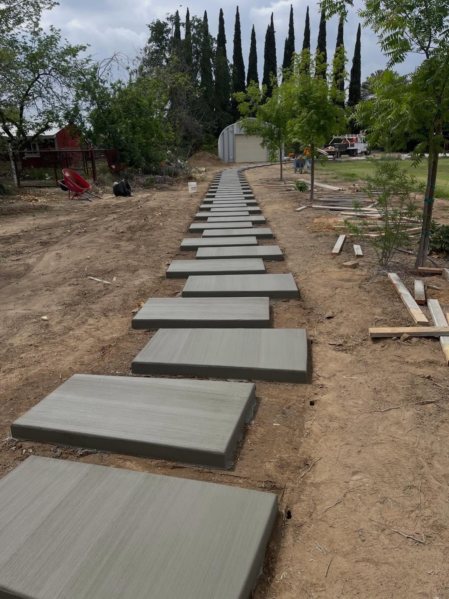 Concrete stepping stones along a dirt path in a garden, lined with trees and a small building in the distance.