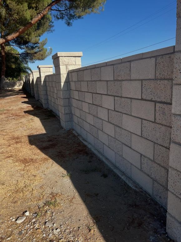Sunlit cinderblock wall along a dirt path beside trees and blue sky