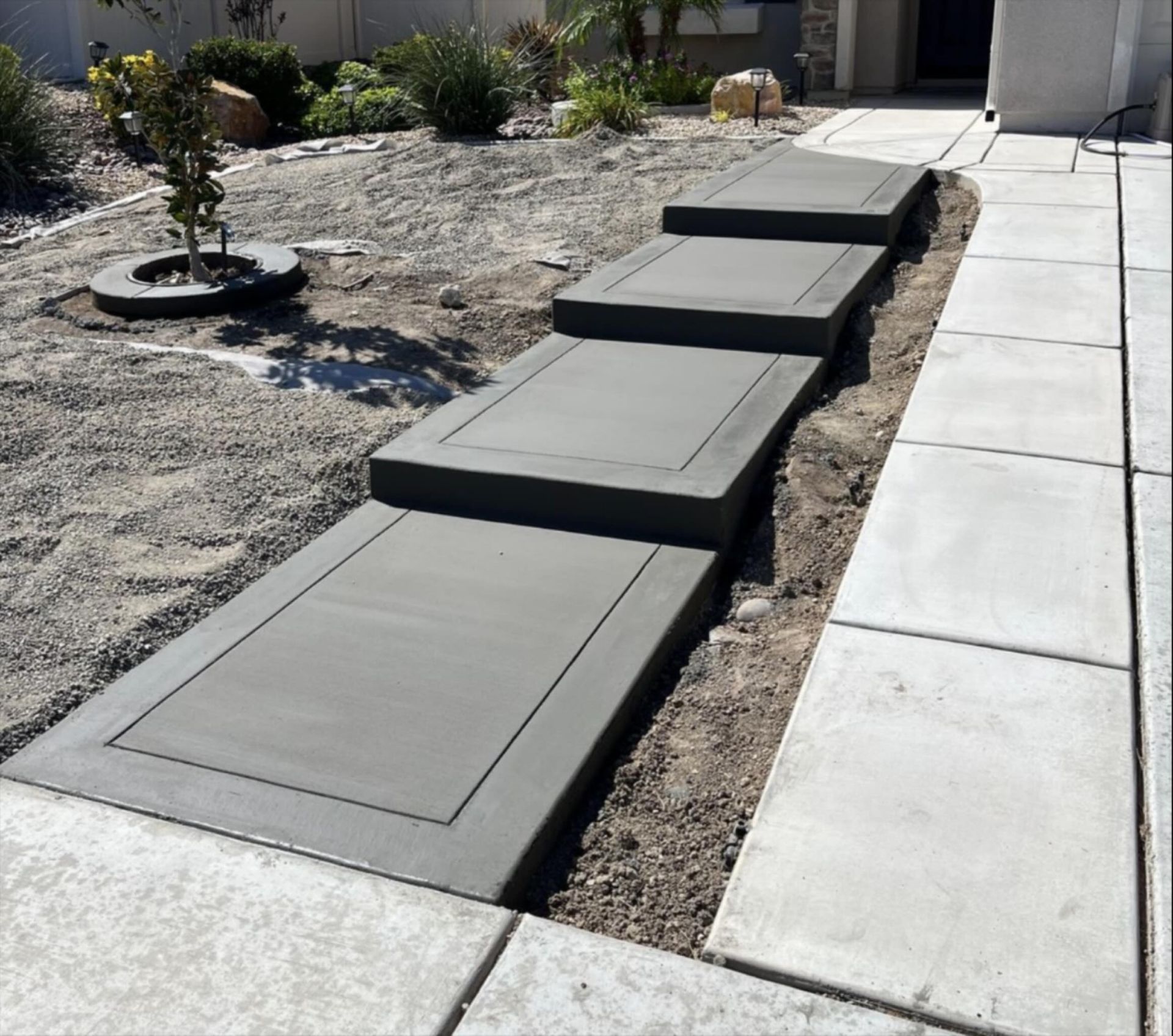Concrete stepping pads along a narrow dirt path beside a sidewalk in a front yard