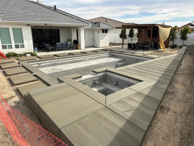 Backyard pool construction with raised spa, beige deck tiles, and a pergola beside a house