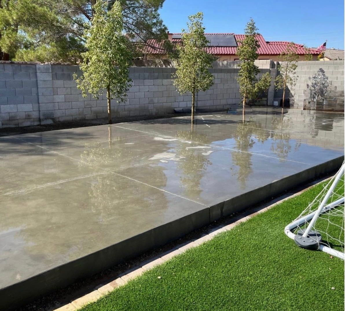 Concrete backyard court with standing water, cinderblock wall, trees, and a soccer goal on grass