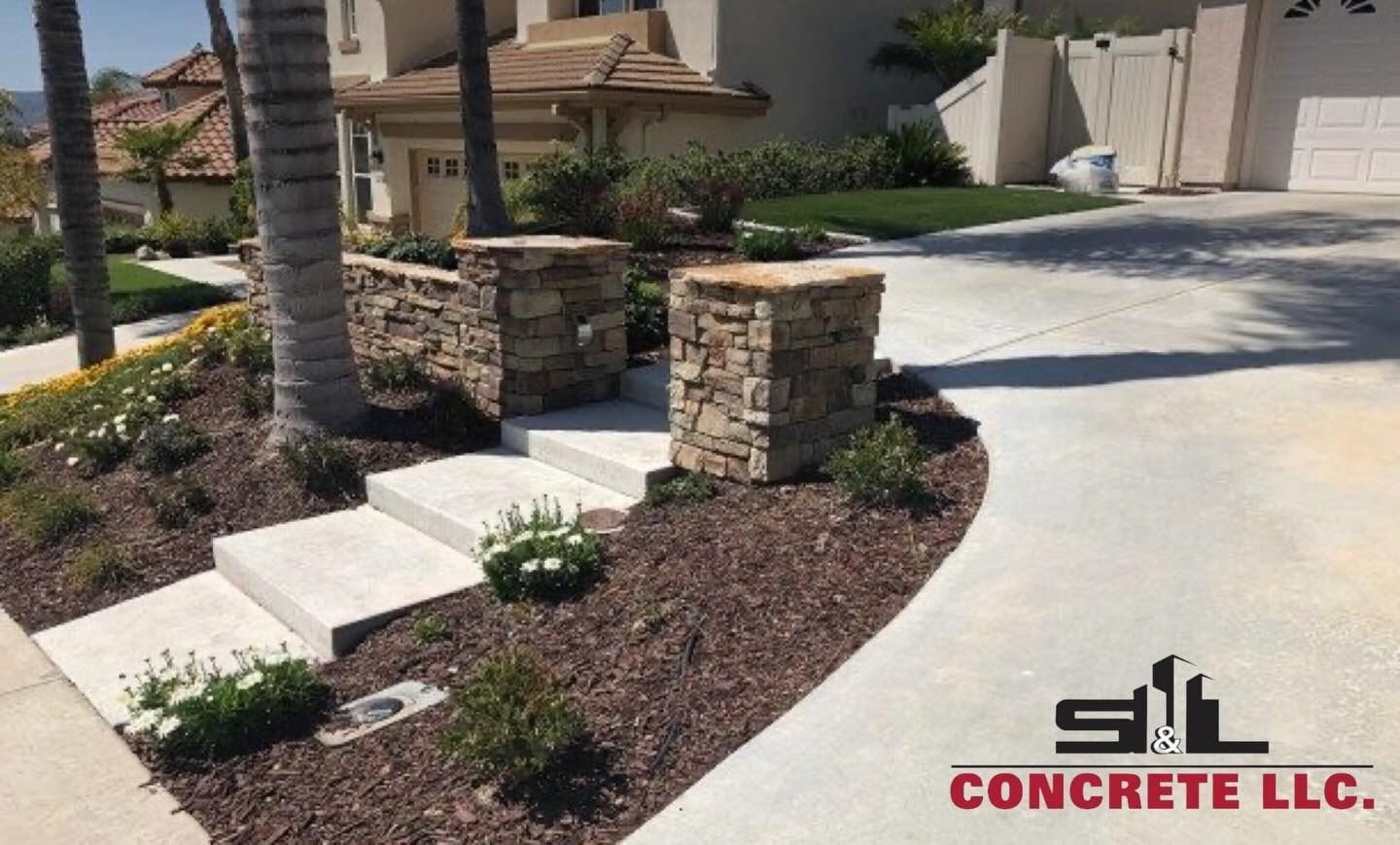 Driveway with decorative stone pillars, concrete steps, and landscaped mulch beds beside a house.
