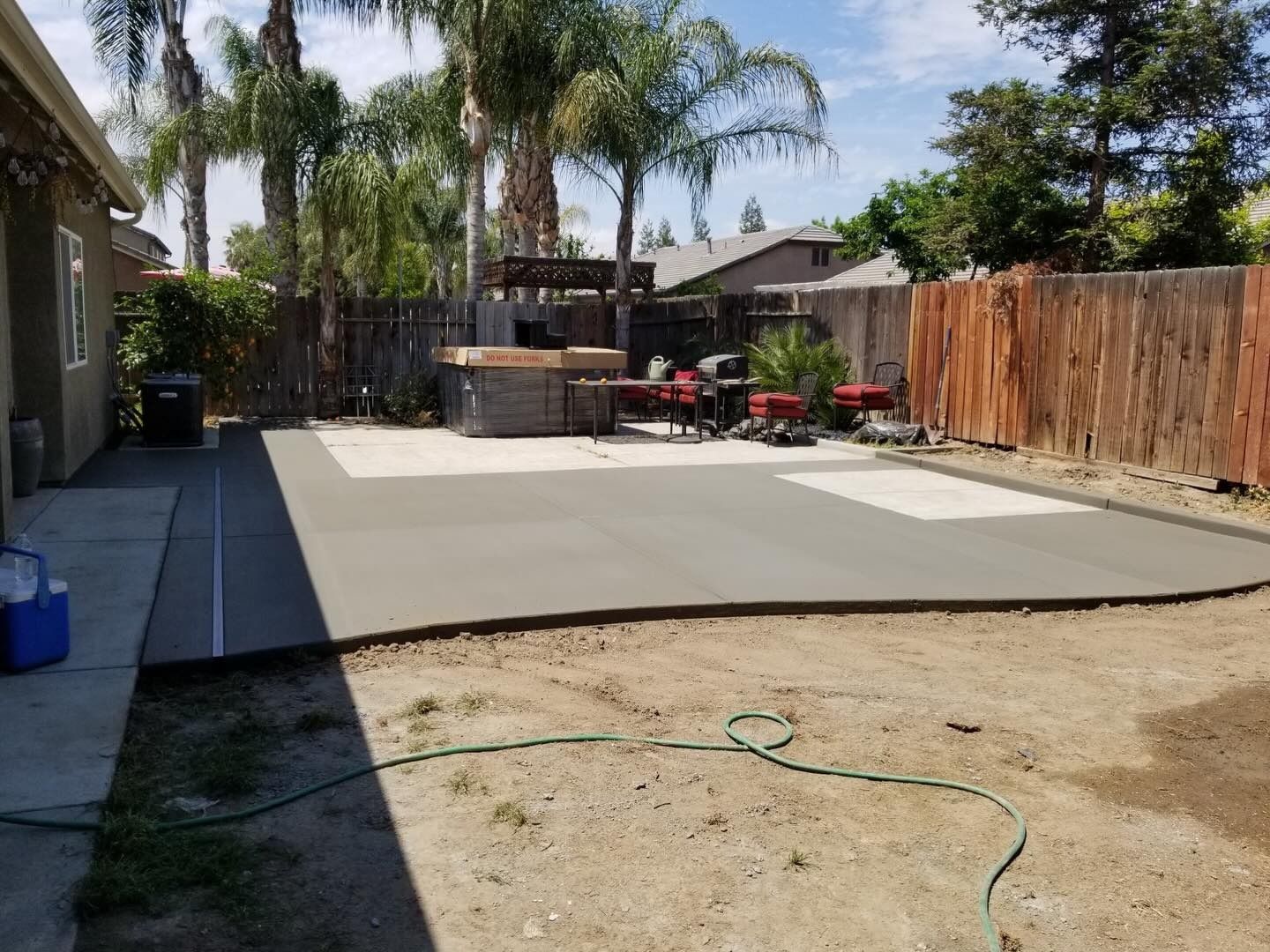 Backyard with a large concrete patio, wooden fence, palm trees, and patio furniture under a bright sky