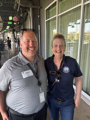 Two people smiling, posing near a window. Man wears a grey shirt, name tag. Woman in blue shirt and jeans, glasses on neckline.