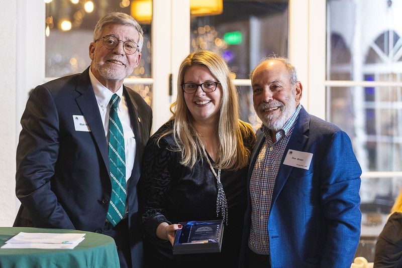 Three people at an event, smiling. Woman holding award, flanked by two men. Background: windows, table.