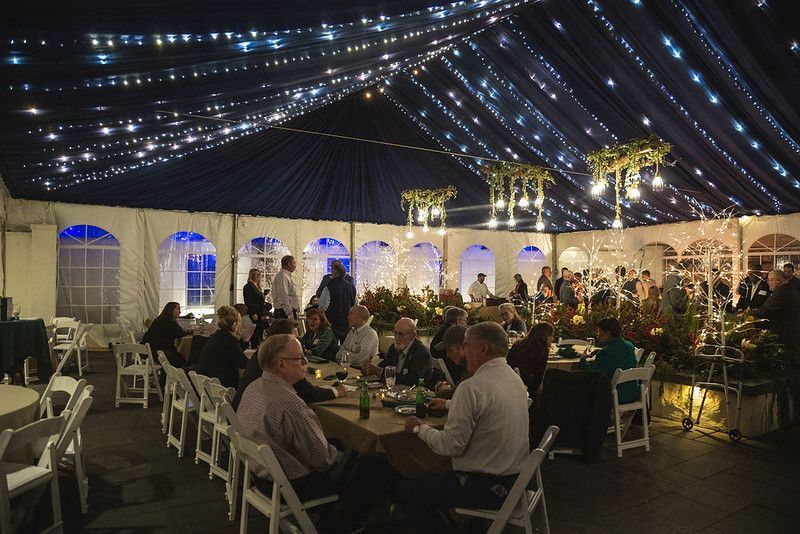 Inside a tent, people dine at tables. String lights adorn the ceiling.