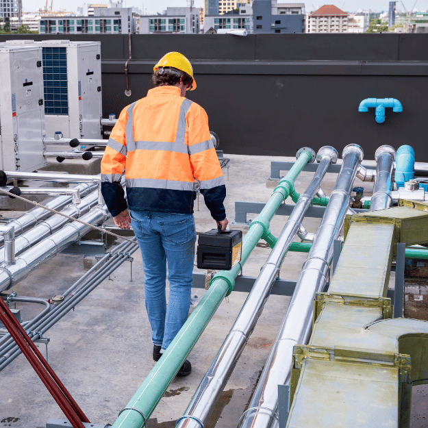 Worker in safety vest and hard hat inspecting rooftop equipment, holding a device.