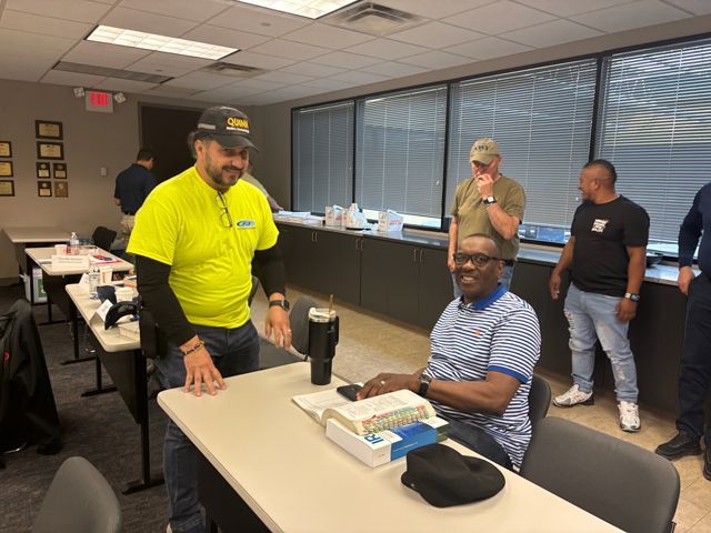 Men in a classroom. One man in yellow shirt smiles, talking to another man in a striped shirt seated at a desk. Other men stand nearby.