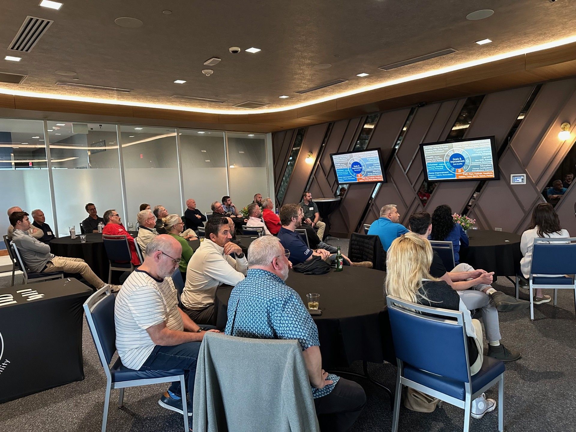 Conference audience seated in a meeting room, facing two wall-mounted presentation screens.