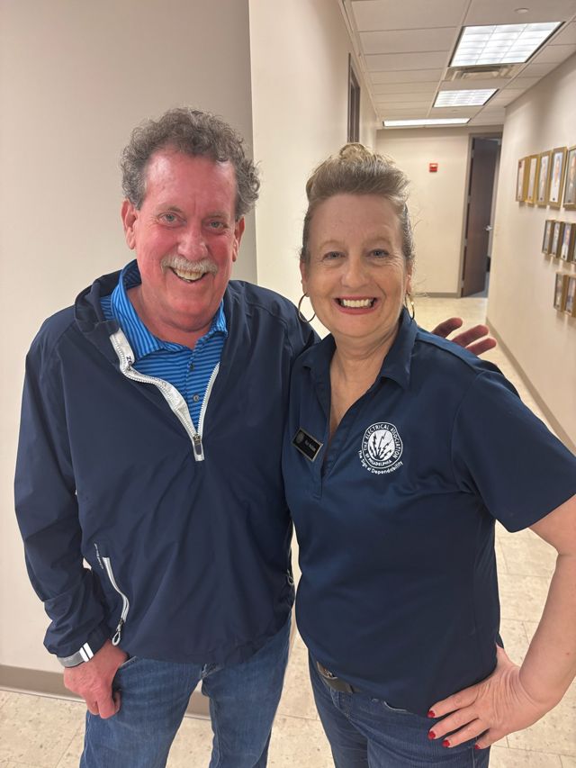 Two smiling people pose in a hallway. The man wears a blue jacket and the woman a blue polo shirt.