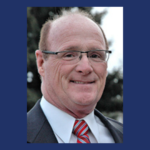 Man in suit and glasses smiles, red tie, outdoor setting.