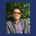 Man wearing glasses and a suit jacket, in front of greenery.