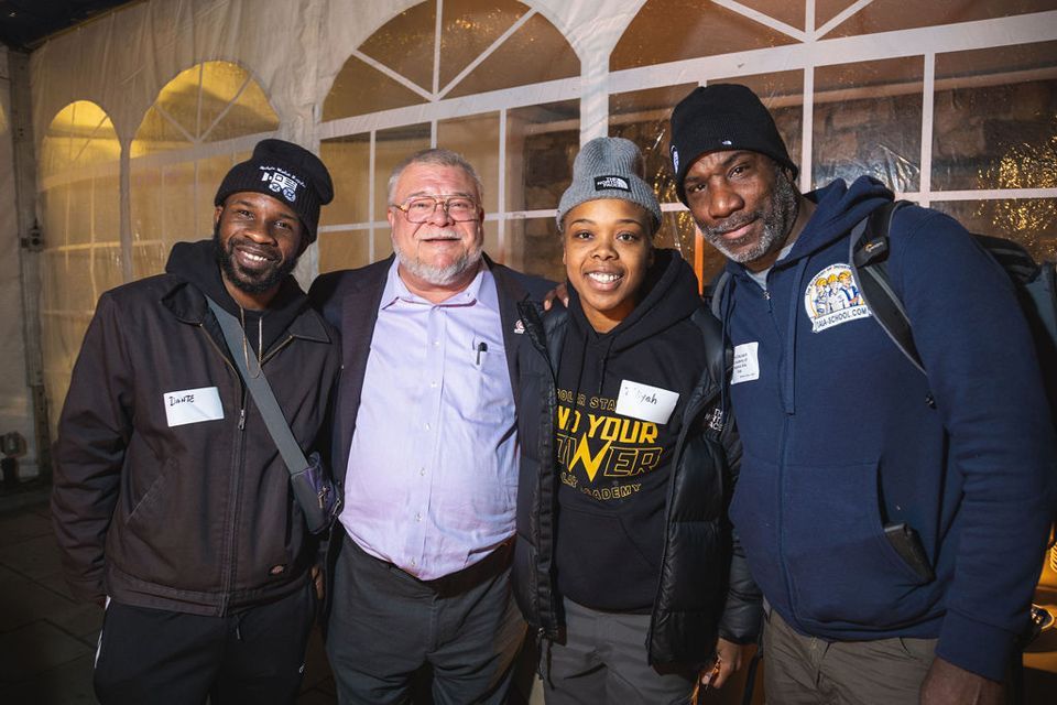 Four people smiling, posing together. Inside a white tent. One person wears a black beanie.