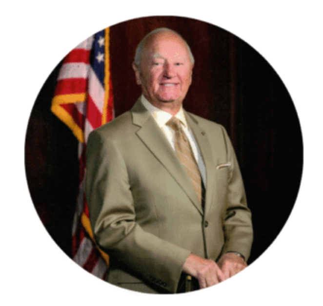 Man in tan suit, smiling, standing in front of an American flag.