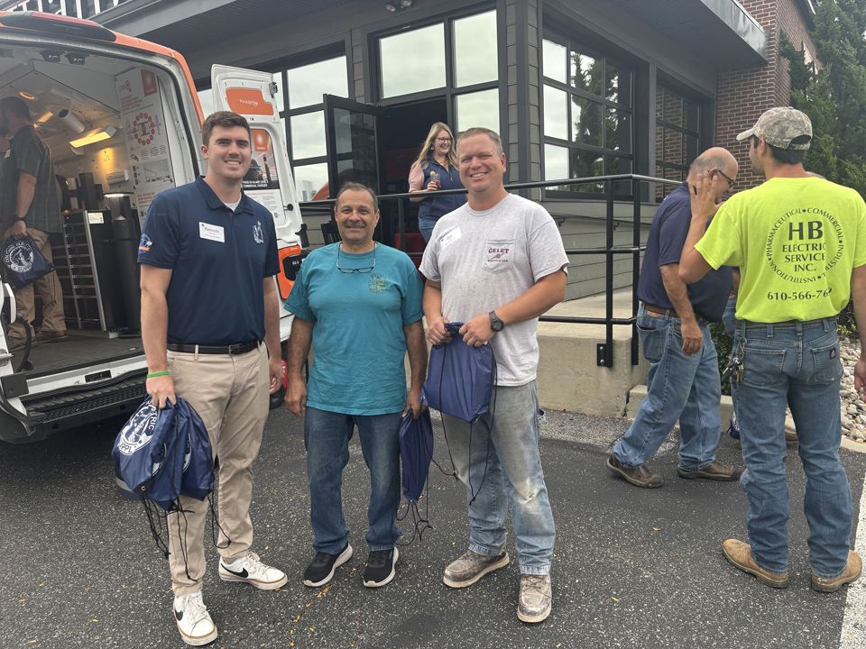 Three men outside building; one in blue shirt, two in t-shirts holding bags. Ambulance in background.