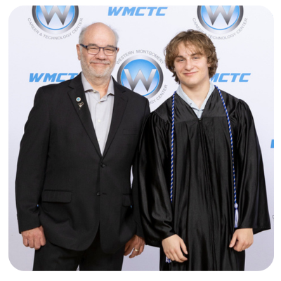 Student in cap and gown receives a check from a man in a suit, on a stage with blue curtains.
