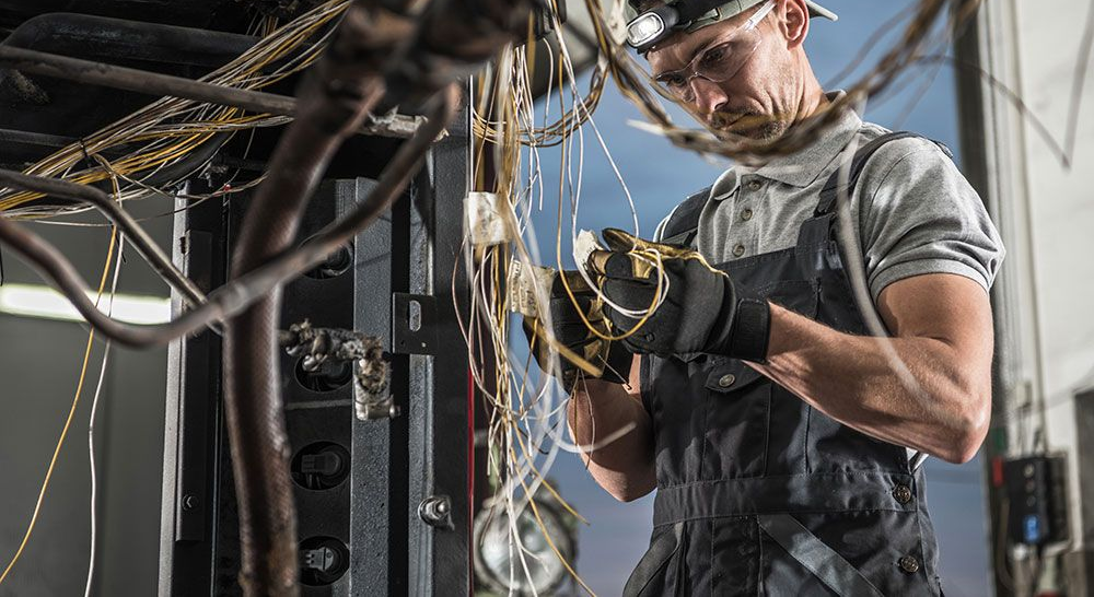 Mechanic in coveralls, headlight, and gloves working on complex wiring in a vehicle.