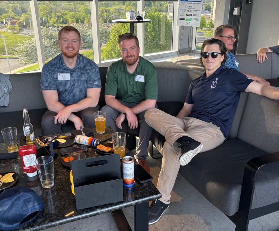 Four men seated on a gray couch at an outdoor table; food, drinks, and a sunny view are visible.