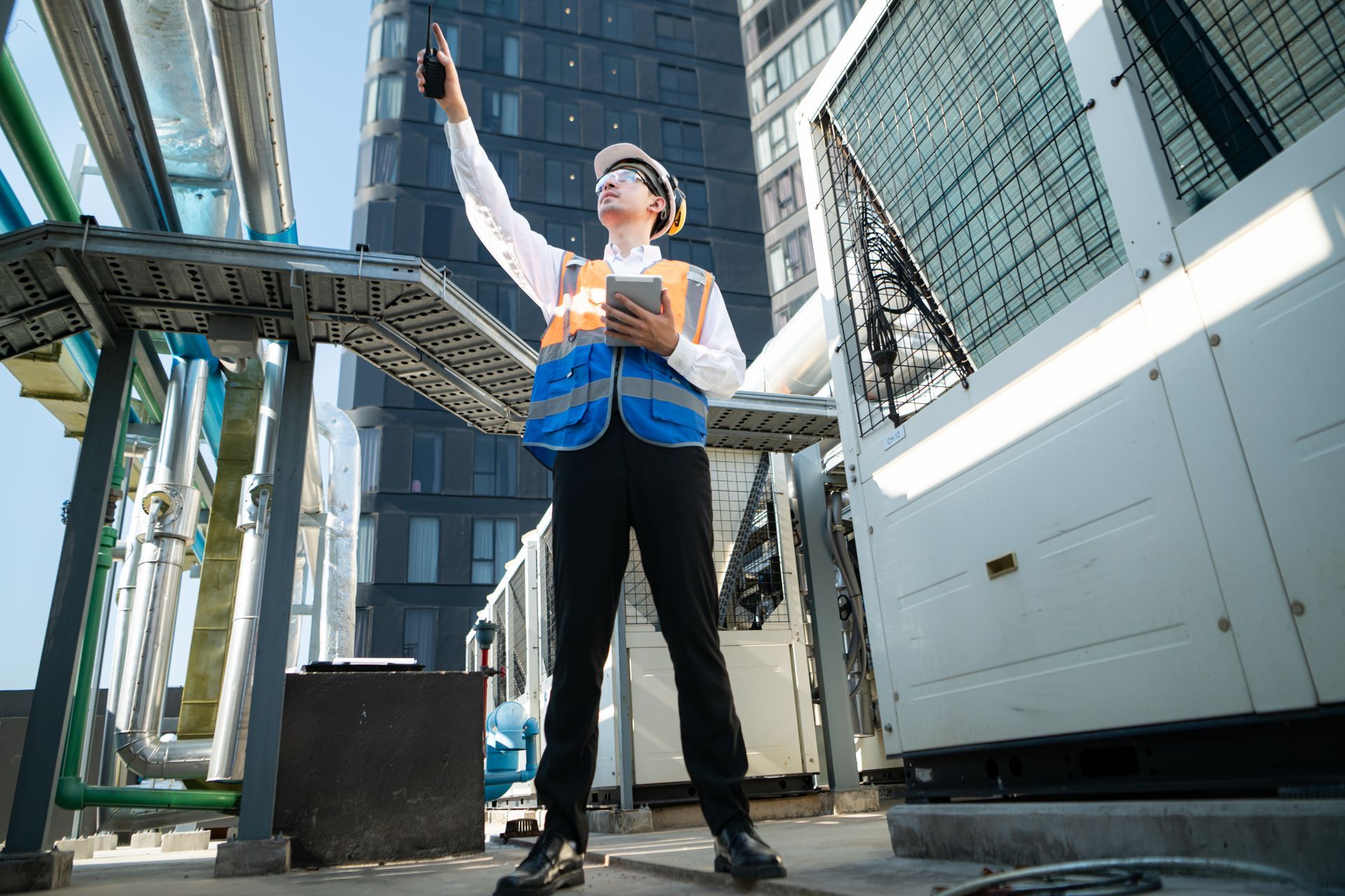 Person in safety vest and hard hat on a rooftop, holding a tablet and device, near machinery and a high-rise building.