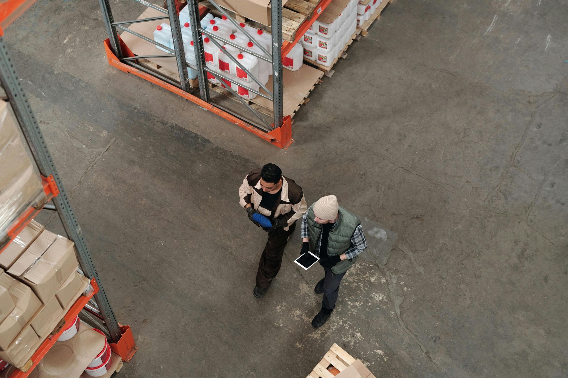Two people walking and talking in a warehouse, near shelves of boxes. One holds a tablet.
