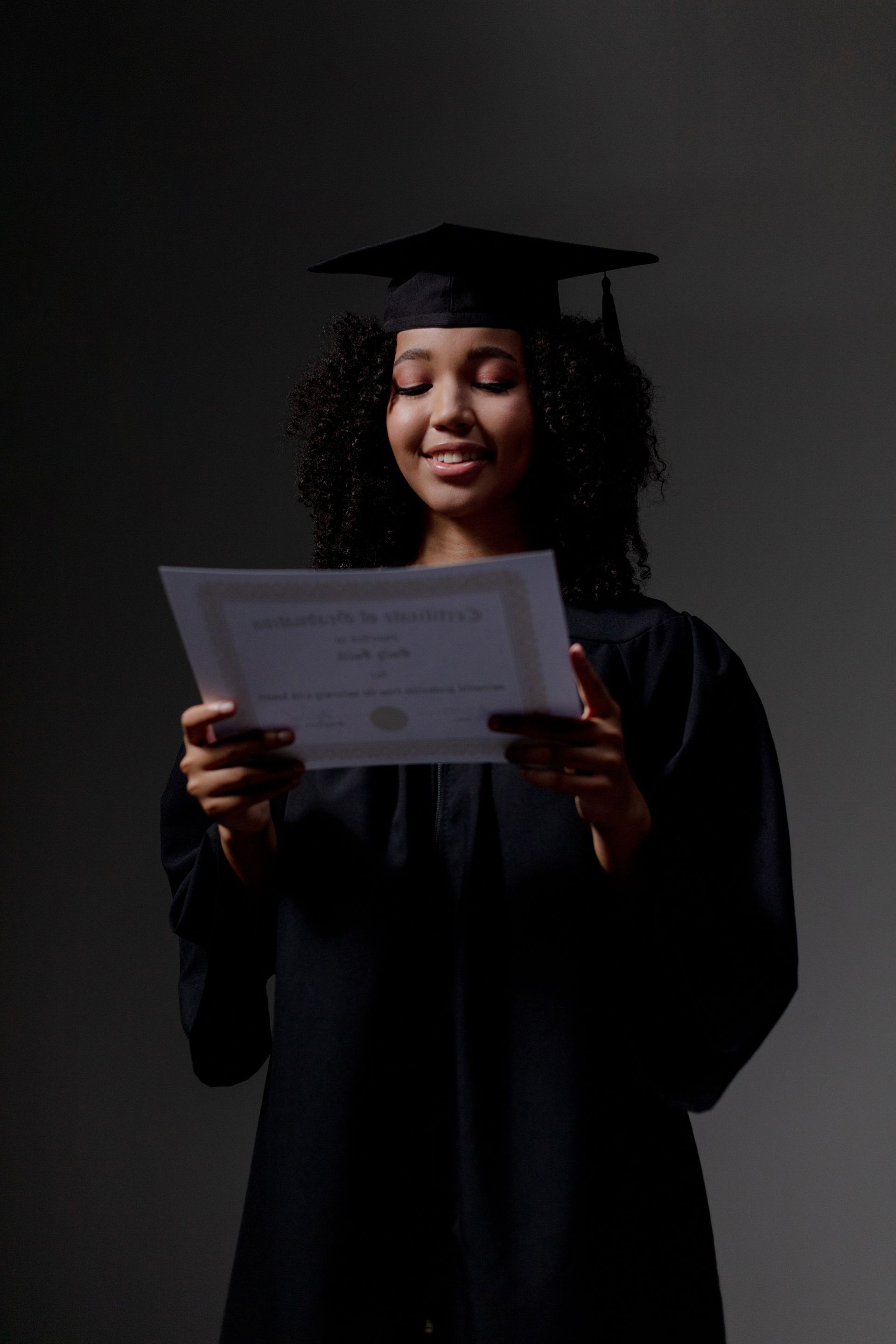 Person in graduation cap and gown holding and reading a certificate against a dark background