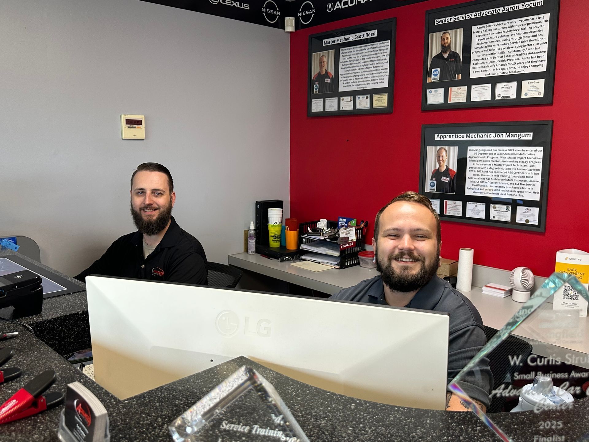 Two men smiling behind a white desk in an office with red and black walls and framed posters | Advanced Car Care Center LLC