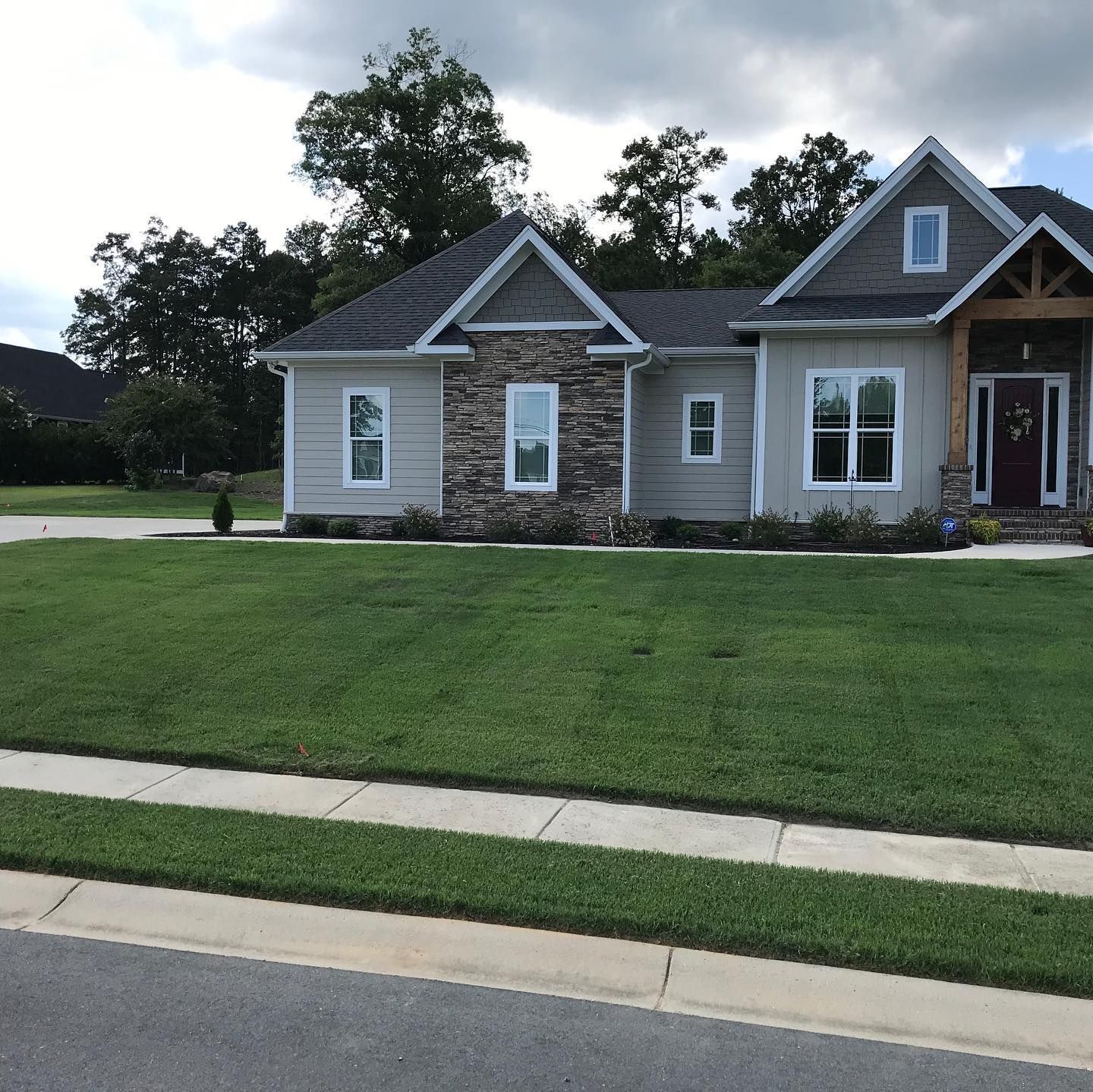 House with beige siding, stone facade, and green lawn. Sidewalk in foreground, trees and sky in background.