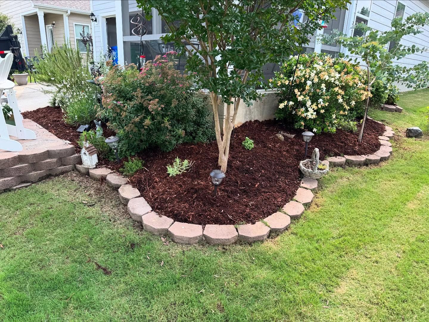 Landscaped garden bed with red brick edging, mulched with plants, tree, and green grass.