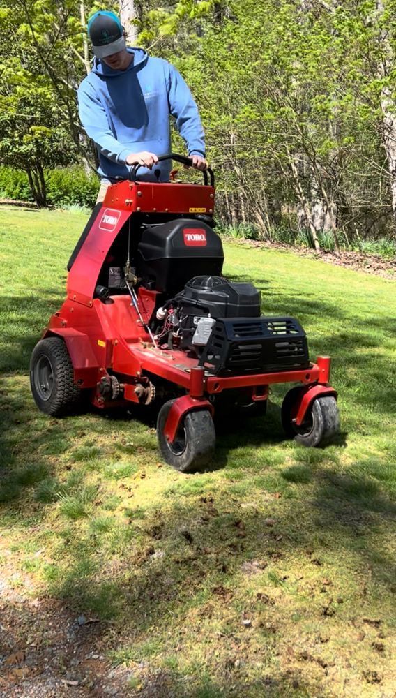 Man operating a red lawn aerator on a green lawn. Trees in the background.