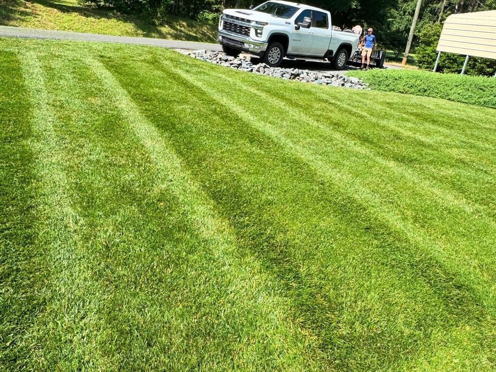 Freshly mowed green lawn with striped pattern, truck parked on gravel, person standing by it on sunny day.