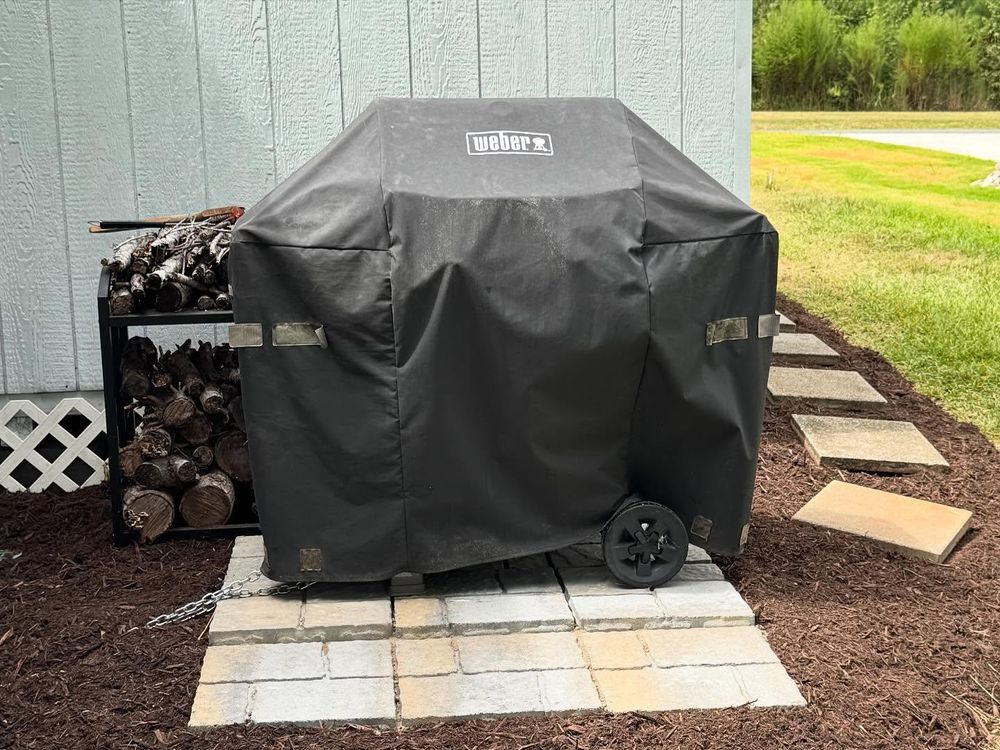 Covered grill on stepping stones next to a wood pile and house.