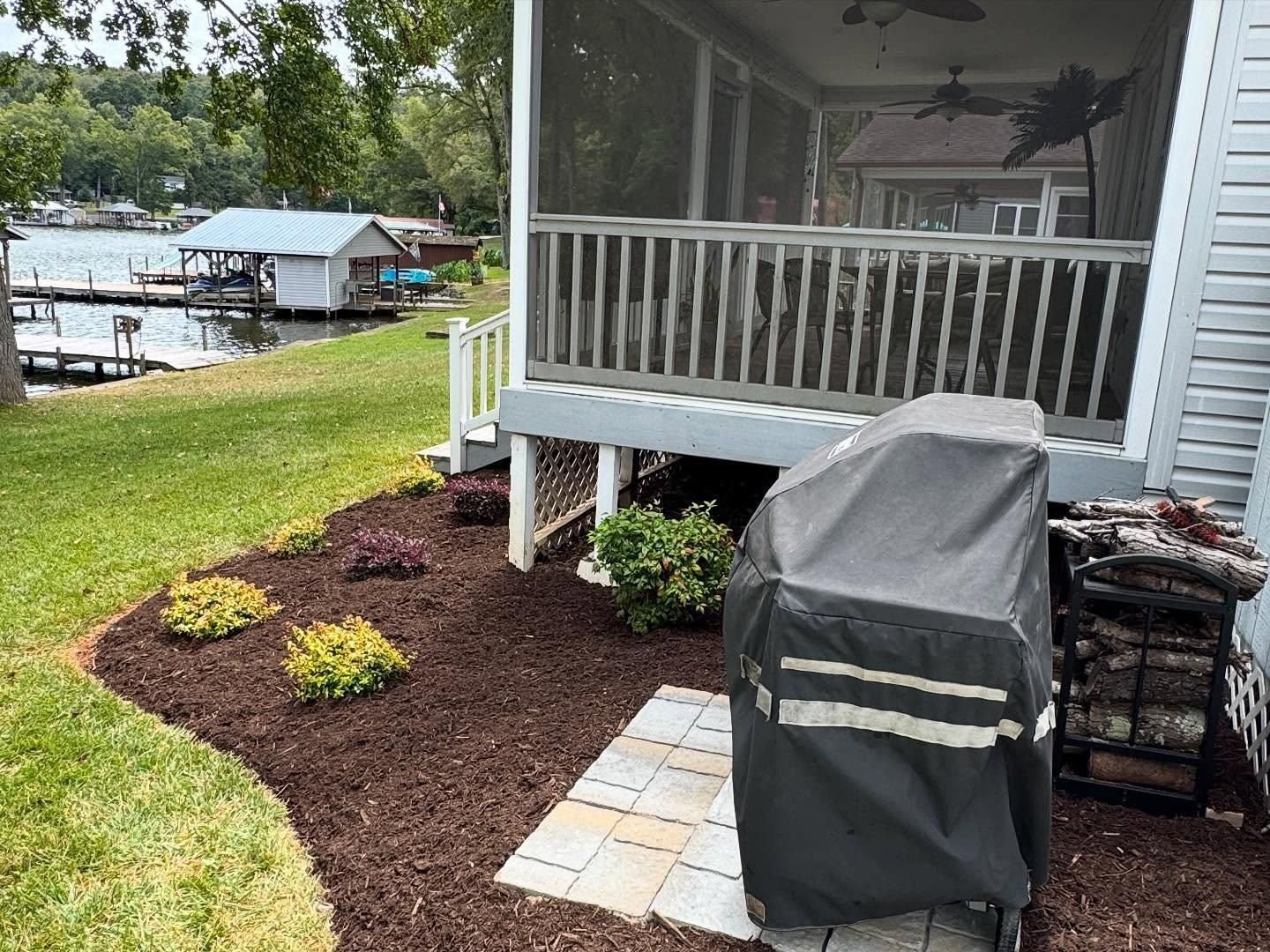 Lakeside porch with grill and firewood. Brown mulch and green grass, with lake and dock visible.