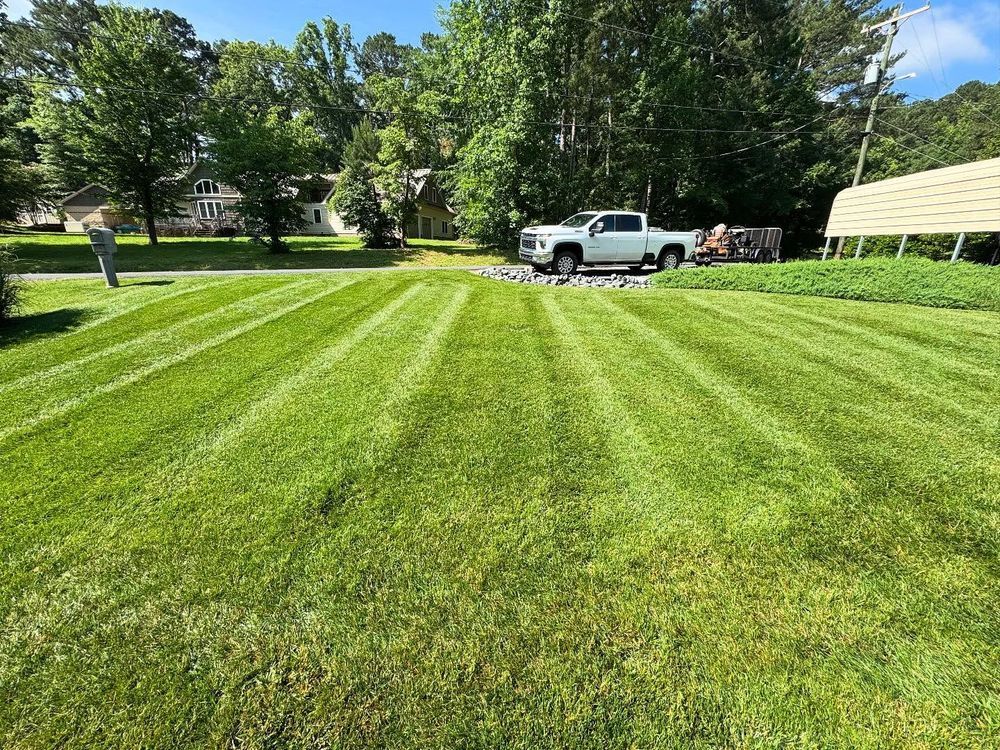 Lawn mowed with distinct stripes, white truck parked on gravel, man standing nearby, trees in background, sunny day.