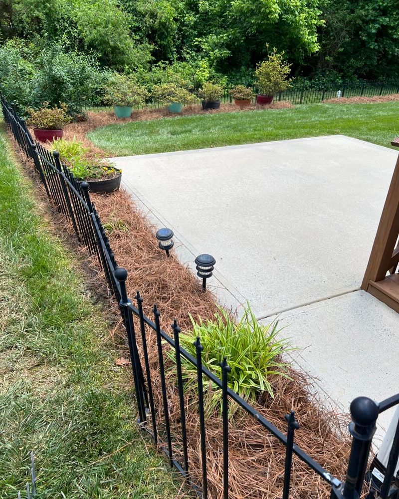Black fence borders a patio with solar lights and potted plants, grass on either side.