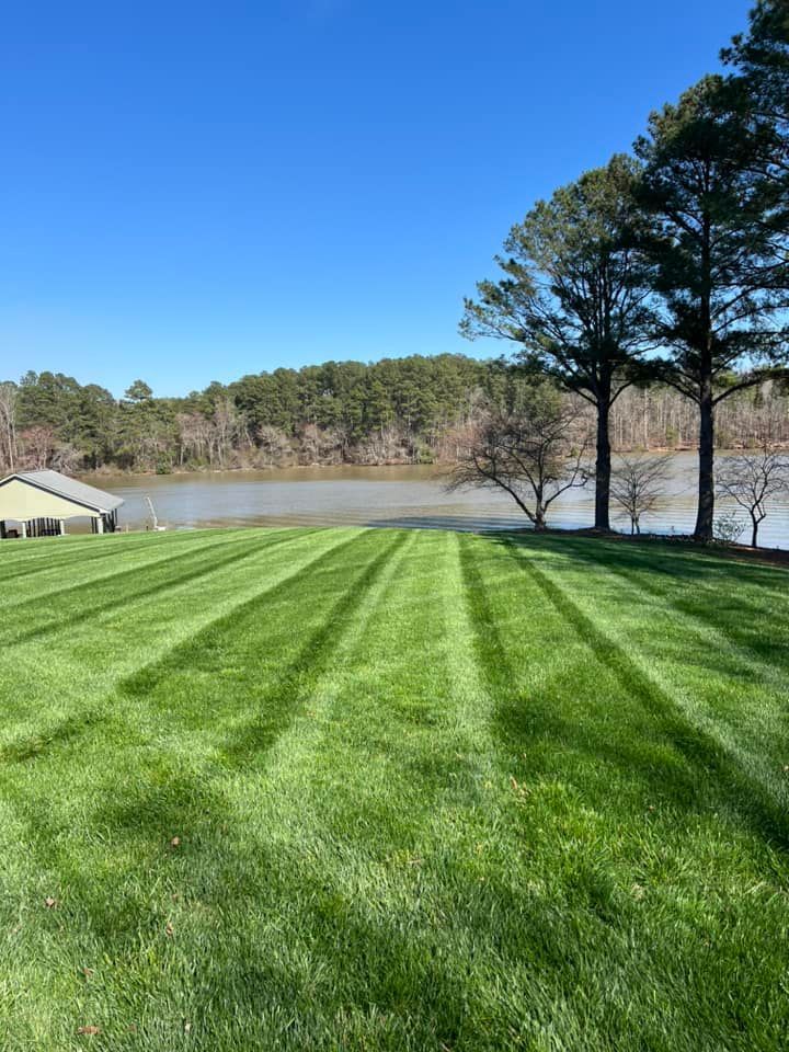 Lush green lawn with mowing stripes leads to a lake, with trees and a building on the left, under a blue sky.