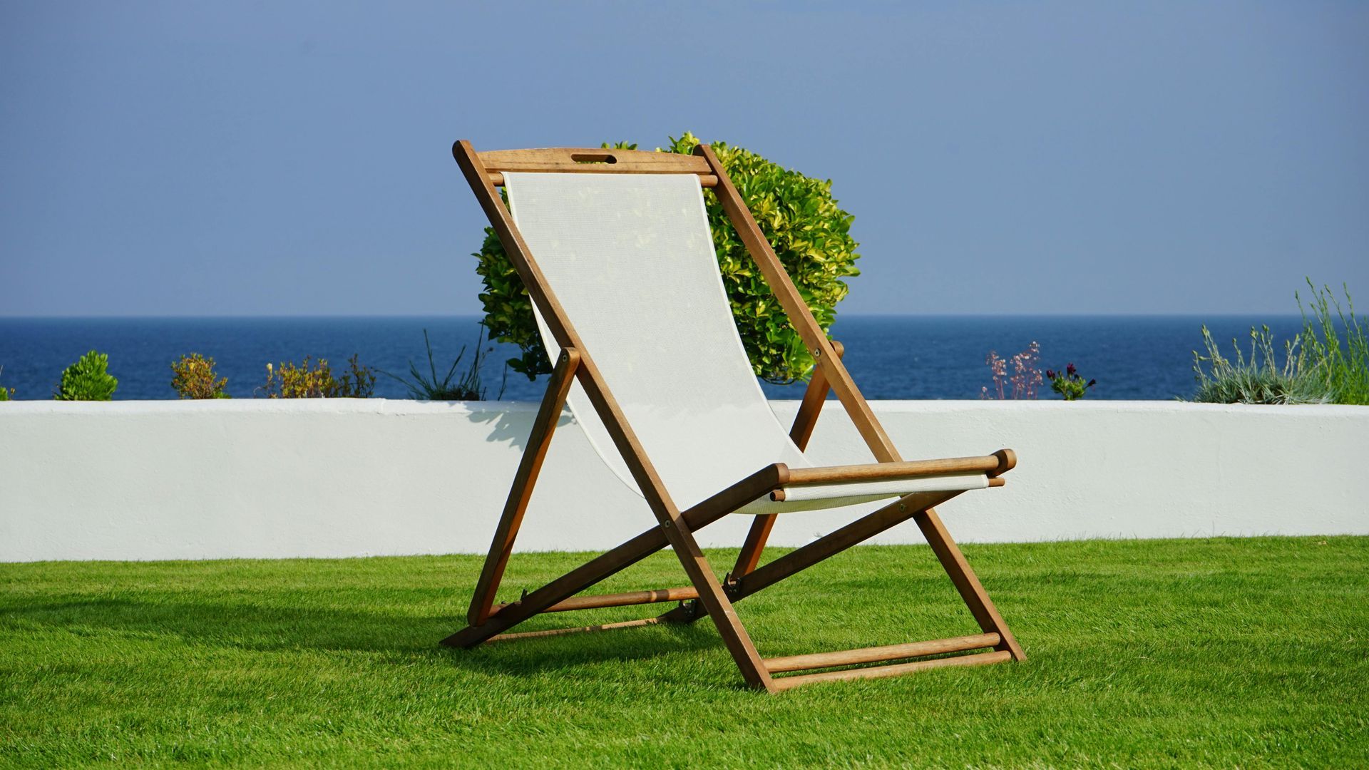 Wooden lounge chair on green lawn facing the ocean, with a white wall and potted greenery behind it.