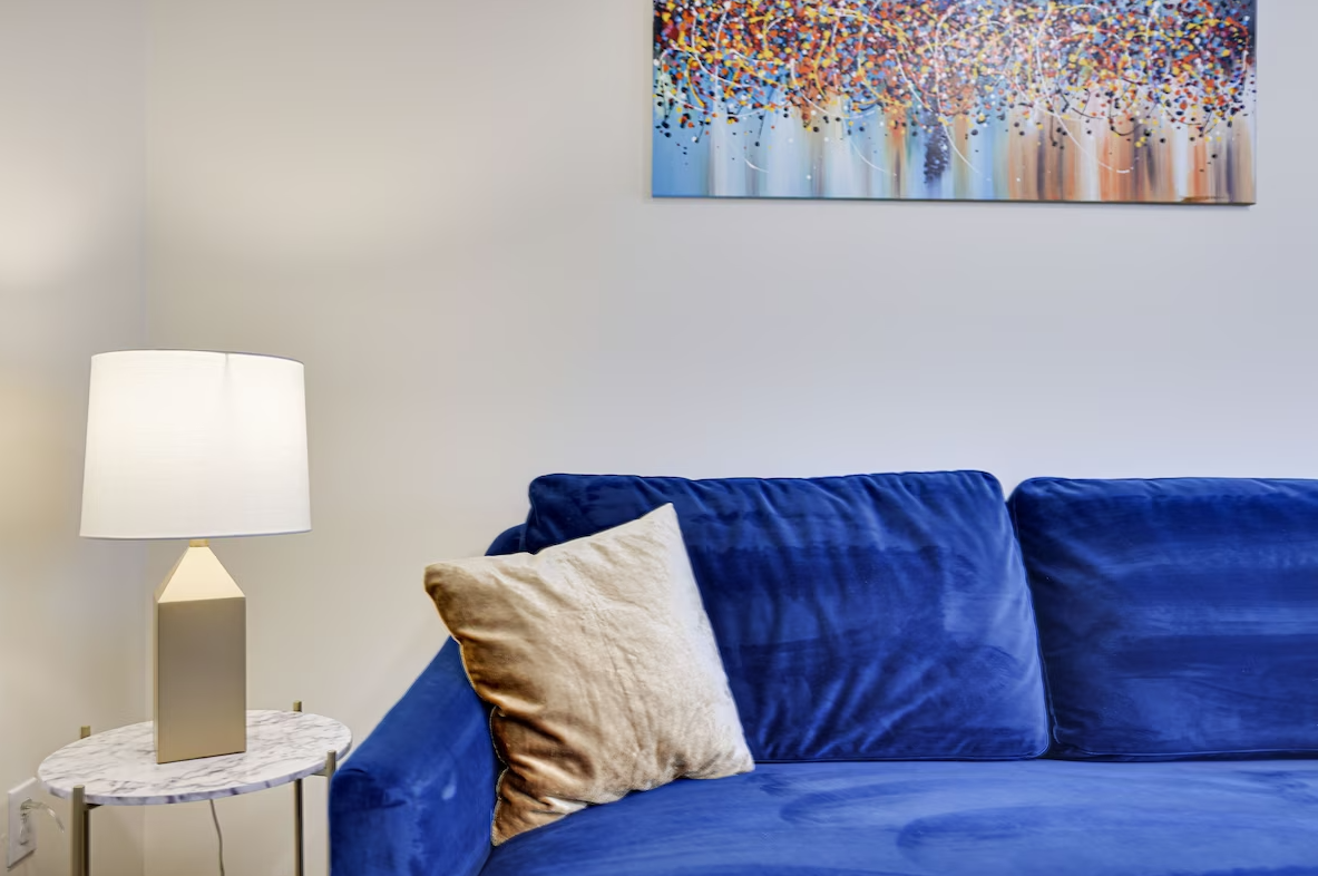 A living room with a blue couch , a lamp , and a painting on the wall.
