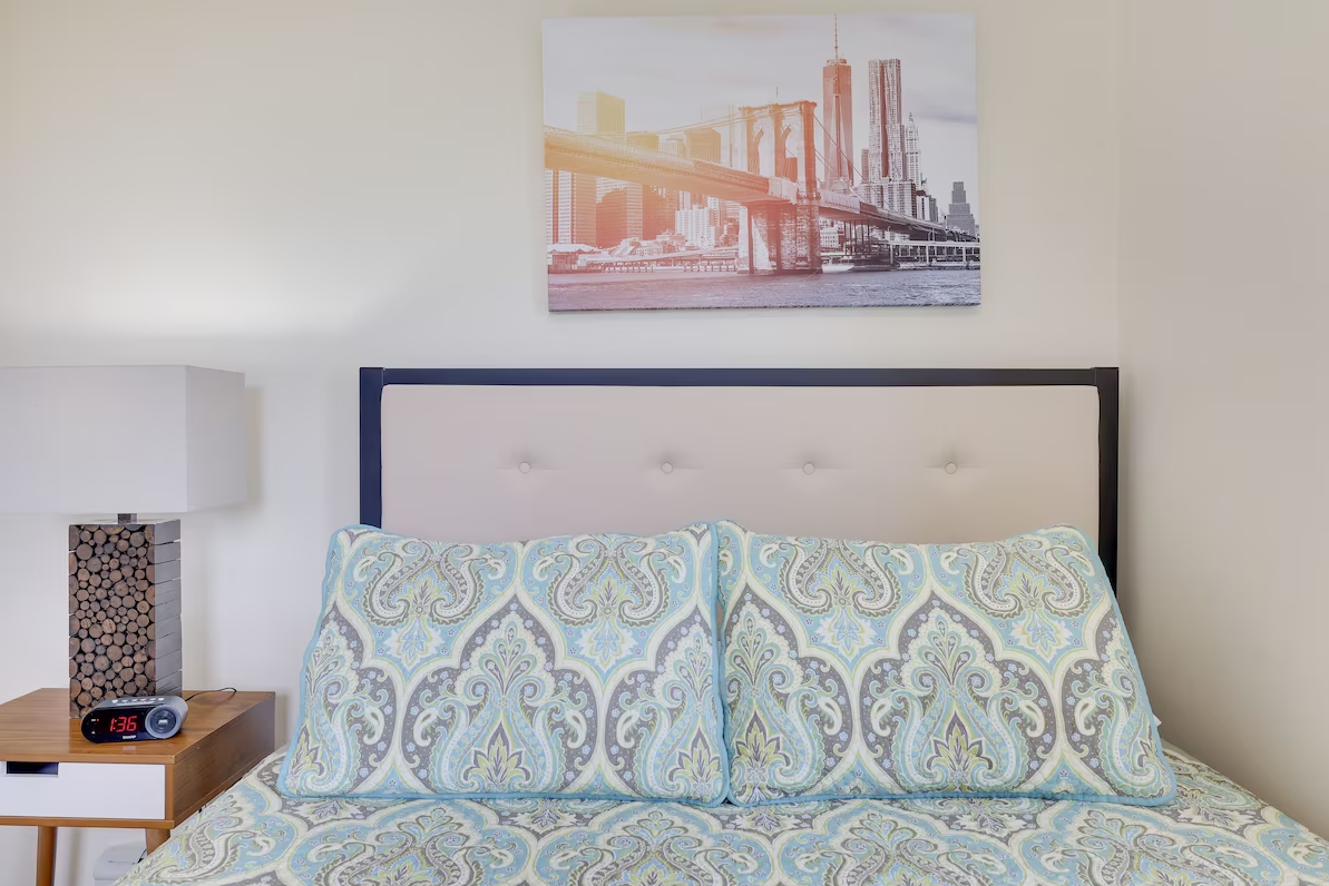 A bedroom with a bed , nightstand , lamp and a painting on the wall above the bed.