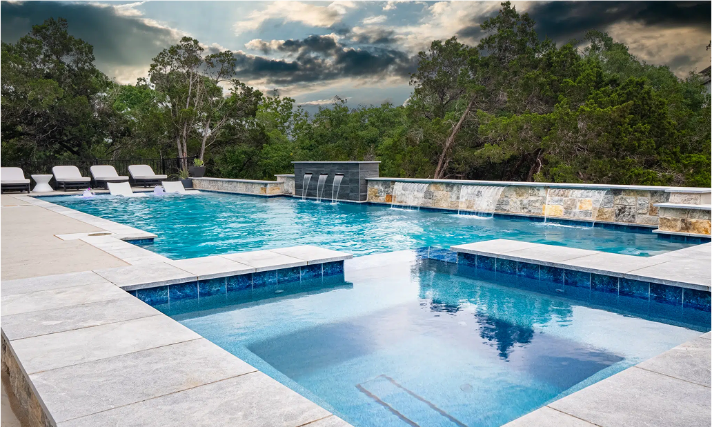 Luxury swimming pool with multiple sections, stone and blue tile, overlooking trees under a cloudy sky.